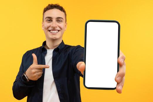 Recommendation. Portrait of excited guy holding big smartphone with white blank screen in hand, showing close to camera and pointing at device. Gadget with empty free space for mock up, banner photo