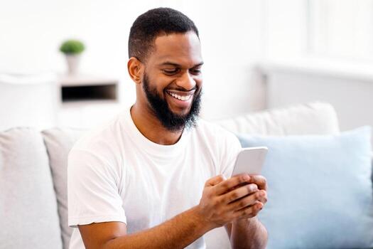Happy Black Man Using Phone Browsing Internet And Networking In Social Media App Sitting On Couch At Home. Cheerful Cellphone User Testing New Application For Smartphone. Mobile Communication Concept photo
