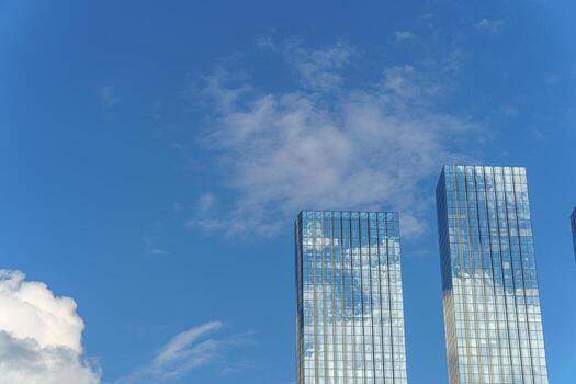 Two glass skyscrapers rise against the blue sky, reflecting clouds and creating a sense of lightness and openness. Their mirrored surfaces form a symmetrical and visually harmonious composition. photo