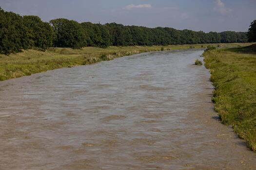 Wide river overflow in urban landscape after heavy rainstorms, with strong current and muddy waters stretching along grassy banks. Floodwaters, river overflow, and environmental change photo