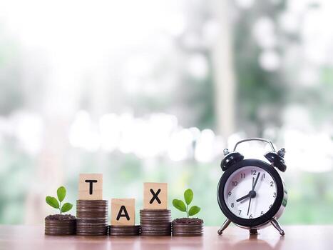 Black alarm clock, Wooden blocks with the word TAX and plants growing up on stack of coins. The concept about saving money and manage time to paying tax in year 2026 photo