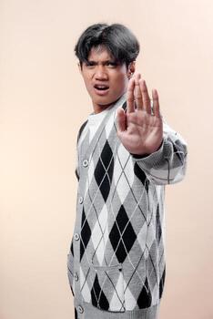Asian man with serious expression showing stop gesture, wearing a grey argyle sweater, hand raised forward, representing rejection, warning, caution, and defensive attitude. photo