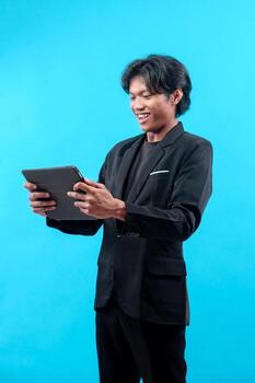 A young Asian man in a black suit is smiling while holding a digital tablet, showing a cheerful and professional expression as he interacts with technology in a modern setting. photo