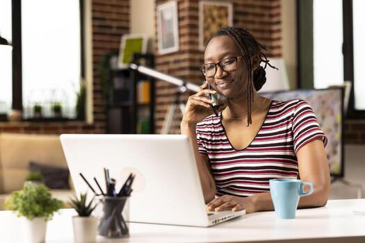 Self employed woman sits at desk, multitasking, speaks with clients on phone and updates data on laptop. Smiling female freelancer uses mobile device while working from home on personal computer. photo