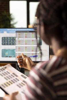 negro hembra persona de libre dedicación estudios analítica en pantalla mientras comparando cifras desde impreso documentos a hogar. de cerca de remoto trabajador con bolígrafo y portapapeles examinando negocio datos en computadora. foto