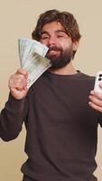 libanesisch jung Mann mit Smartphone Telefon Mit freundlichen Grüßen Jubel gewinnen, Empfang Geld Dollar Kasse Banknoten, Erfolg Lotterie Jackpot Glück, Einkommen Gehalt Lohn. indisch Hindu Kerl auf Studio Beige Hintergrund. video