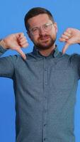Dislike. Upset unhappy Caucasian man showing thumbs down sign gesture, expressing discontent, disapproval, dissatisfied negative feedback. Handsome attractive guy indoors isolated on blue background video