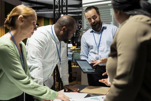 Multiethnic project team shares analytics reports during advisory board briefing late at night, reviewing business intel and preparing metrics for upcoming presentation with shareholders. photo