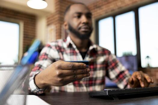 Closeup of male hands holding credit card and making secure online payment on computer. Black man shops digitally using banking website, typing information on desktop pc for convenient checkout. photo