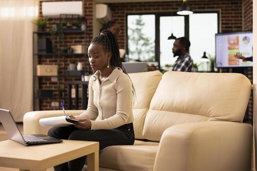 Professional woman reviewing company documents and uses laptop for data analysis in startup office. Female manager working on business project, sits at couch, using both digital and printed resources. photo