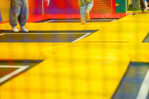 Bright yellow and black trampolines are visible in a play area. The surface is padded, and colorful netting surrounds the space. Soft lighting illuminates the area. photo