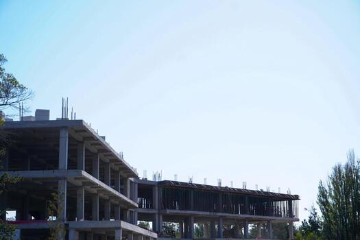 A concrete building structure is partially completed with exposed beams and columns. The sky is clear and bright, illuminating the construction site. photo