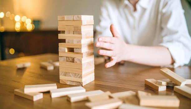 Wooden block game tower with scattered pieces on table and person hand playing, creating tense and focused atmosphere in warm indoor setting photo