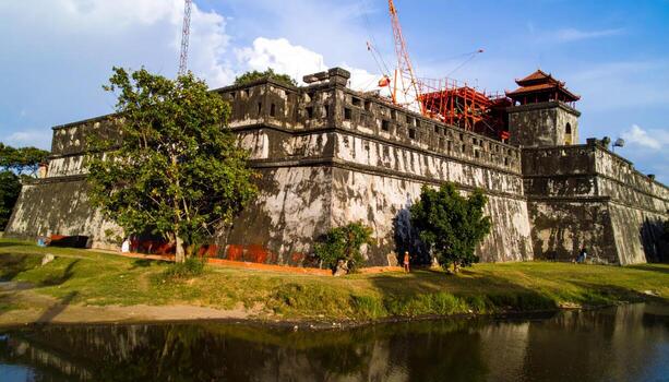 Ancient stone fortress with weathered walls and traditional roof structures beside calm river under partly cloudy sky, surrounded by green grass and trees in daylight photo