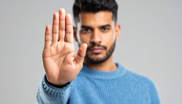 Serious man in blue sweater holding hand up in stop gesture, focusing on palm, expressing refusal or warning, isolated on gray background photo