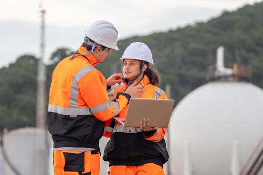 Team of Engineers Collaborating at Oil and Gas Storage Facility, Industrial Workers Using Laptop to Monitor Operations at Site, Engineers Communicating and Supervising Work at Petroleum Storage Tanks photo