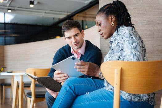 A diverse business team collaborating on a tablet in a modern office or cafe setting, Two colleagues, a man and a woman, meeting and reviewing a digital document and notebook together photo