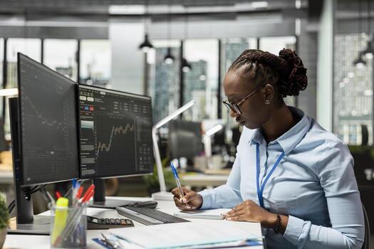 Risk analyst in office documents strategies while observing stock candle patterns on screen. Brokerage company worker reviews stock market shares volatility, recording insights in notepad photo