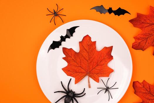 White plate with autumn maple leafs, spiders and black bats on bright orange background. Halloween party and table setting concept. Flatlay. Top view. Copy space. photo