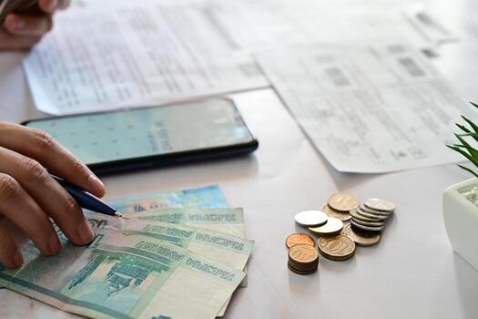 A close-up shot showcases a hand holding a pen over a document next to a smartphone displaying a calculator app, with Russian currency and coins nearby. photo