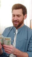 jung Mann beim Zuhause halten ein Stapel von Dollar Kasse Banknoten, Zählen Geld mit Aufregung. kaukasisch Kerl auf Sofa lächelt, Gefühl glücklich Über ein groß Gehaltsscheck Lotterie Sieg oder unerwartet finanziell Erfolg video
