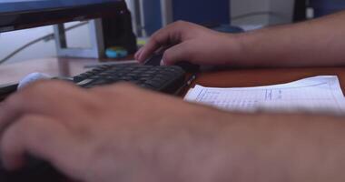 un' angolo basso, medio tiro di un' di persona mani e avambracci Lavorando a un' scrivania, digitando su un' tastiera e utilizzando un' topo. un' bloc notes con un' simile a un foglio di calcolo griglia è visibile Il prossimo per il tastiera del computer. video