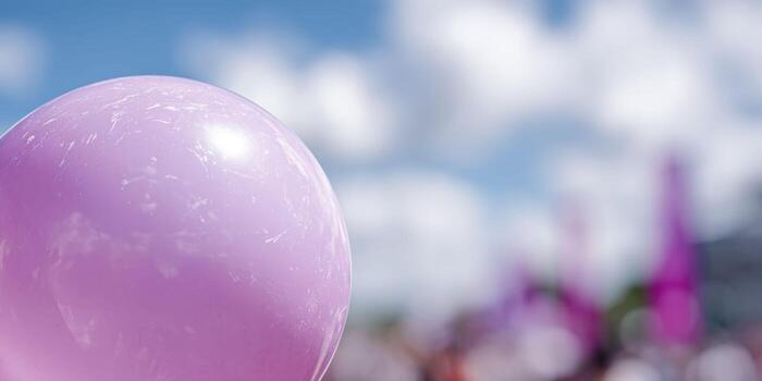 A macro view of a pink balloon surface, capturing its glossy texture and slight imperfections under bright daylight, with a blurred festival banner and cheering crowd in the background. photo