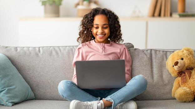 Communication and technology. Smiling black teen girl sitting on sofa and using laptop computer, doing homework, browsing internet, watching or movie, typing on keyboard and smiling photo