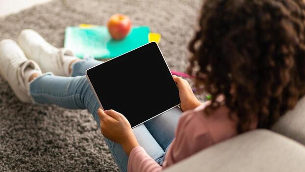 African american curly schoolgirl using digital tablet with blank screen, using mobile application or useful website while resting after study at home. Empty space on device for mockup photo