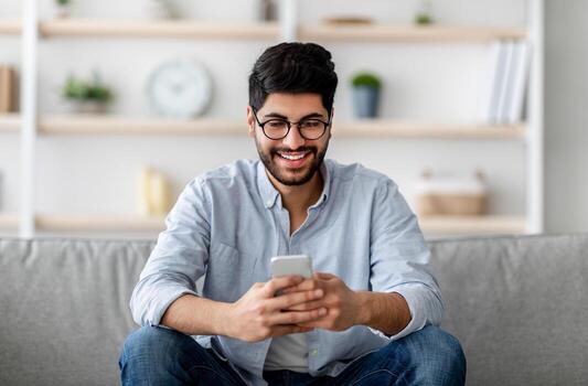 Happy arab guy in glasses sitting with smartphone in hands and messaging or browsing social networks, relaxing with cellphone on couch, checking new app, copy space photo