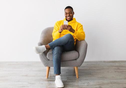 New Application. African American Guy Using Smartphone With Great Mobile App Texting Sitting In Chair Over Gray Wall Background. Man With Cellphone Chatting And Networking In Social Media photo