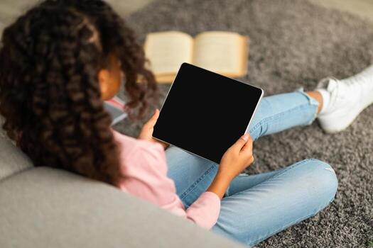 E-learning concept. African american schoolgirl using digital tablet with empty screen, surfing useful website or watching online lesson while sitting on floor with books. Mockup for your design photo