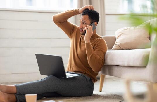 Confused Discontented Arabic Guy Talking On Cell Phone Using Pc Having Problem With Computer Or Internet Connection At Home. Displeased Male Customer Calling To Hotline Service And Complaining photo