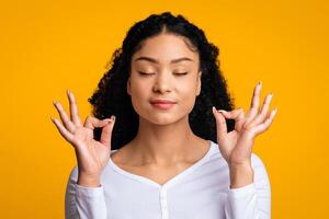 zen. relajado joven africano americano dama meditando con cerrado ojos, satisfecho negro dama acuerdo manos en mudra gesto, en pie aislado terminado amarillo fondo, panorama con Copiar espacio foto