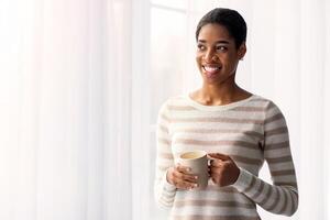 contento sonriente africano americano mujer Bebiendo café mientras en pie cerca ventana a hogar, alegre negro milenario hembra participación taza con caliente bebida y mirando lejos, disfrutando fin de semana, Copiar espacio foto