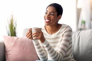 fin de semana ocio. alegre joven negro ama de casa Bebiendo café a hogar mientras relajante en cómodo sofá en vivo habitación, sonriente africano americano mujer participación taza con caliente bebida y mirando lejos foto
