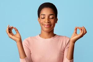 mente armonía. calma relajado negro mujer meditando con cerrado ojos, satisfecho africano americano dama practicando yoga, acuerdo manos en mudra gesto, en pie aislado terminado azul fondo, gratis espacio foto