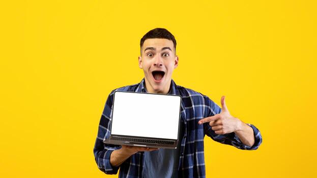 Excited Caucasian man pointing at empty laptop screen with mockup for website design on orange studio background. Emotional young guy showing computer with space for your ad photo