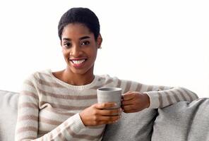 fin de semana pasatiempo. retrato de contento negro hermosa dama con taza de café relajante en sofá, joven africano hembra disfrutando caliente bebida mientras descansando en vivo habitación y sonriente a cámara, Copiar espacio foto