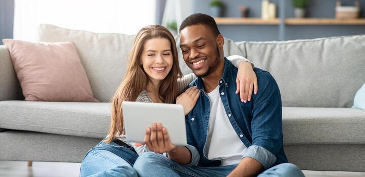 Young Interracial Spouses Using Digital Tablet Together While Resting At Home, Happy Multicultural Couple Watching Movie Or Browsing Internet On Tab Computer While Relaxing On Floor In Living Room photo