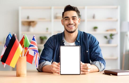 Handsome middle-eastern young man showing pad with blank screen and smiling, sitting at workdesk with notepad and flags of different countries, studying foreign languages online, mockup, copy space photo