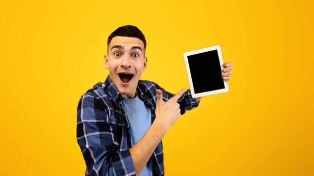 emocionado hipster chico señalando a tableta con blanco pantalla en naranja fondo, espacio para solicitud o sitio web diseño. positivo milenario hombre demostración toque almohadilla con Bosquejo para tu anuncio foto