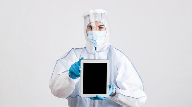 Doctor in protective hazmat suit holding tablet computer with empty screen on light studio background, mockup for mobile app or website design. Medical worker showing touch pad with blank space photo