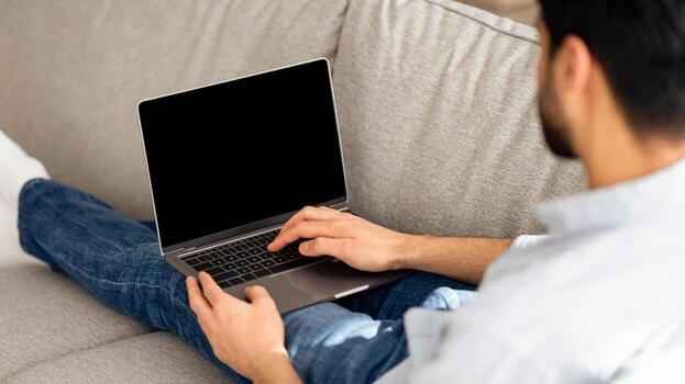 Young arab guy using modern laptop with empty black screen at home, mockup. Unrecognizable millennial man reclining on couch and using notebook with blank display, over shoulder shot photo