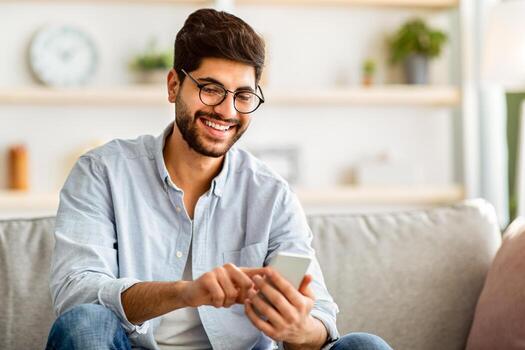 comprobación nuevo aplicación contento árabe hombre utilizando teléfono inteligente, mensajería con amigos, disfrutando móvil comunicación o hojeada social redes, relajante con teléfono en confortable sofá a hogar foto