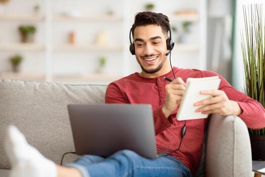 Cheerful middle-eastern young man having online class while staying home, sitting on couch with modern laptop, using headset, looking at notebook screen and smiling while taking notes, copy space photo