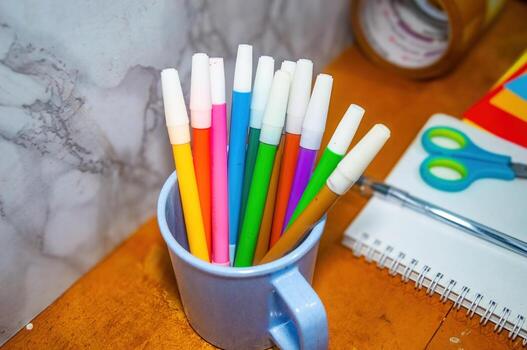 Overhead shot displays a cup filled with colorful markers, alongside a notebook and scissors, set on a wooden table, perfect for illustrating creative workspace photo