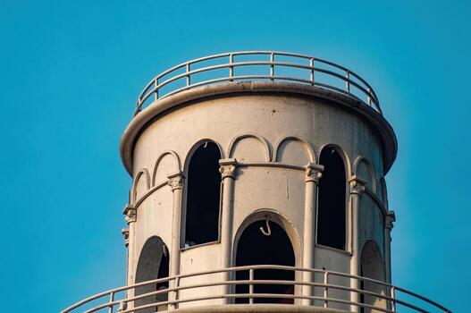 Close up of a vintage lighthouse tower against a clear blue sky, featuring architectural details like arched windows and metal railings. The structure shows signs of age and weathering. photo