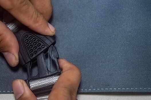 Close up on hands fastening a black plastic buckle on a blue fabric bag. The buckle features a grid pattern, and the bag has a visible stitched edge, showcasing detail and utility photo
