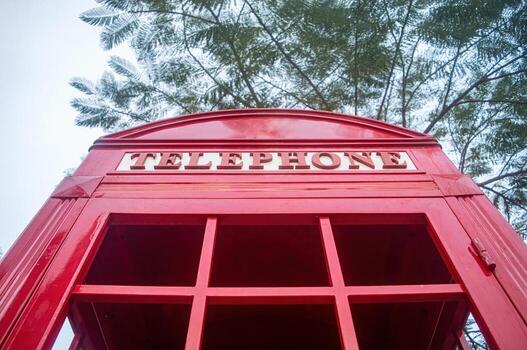 Close up of a red british telephone booth with the word telephone on top, set against a backdrop of tree branches. It evokes a sense of nostalgia and british culture. photo